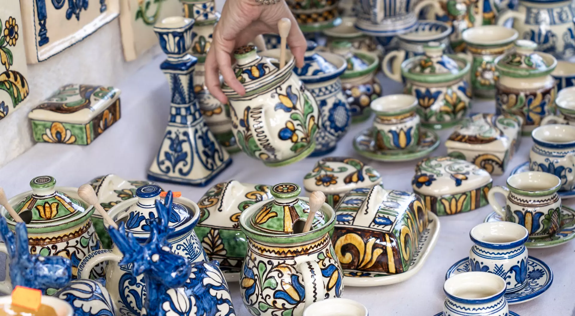 A hand reaches for a colorful, ornate pottery jar among various other decorated ceramics on a display table.