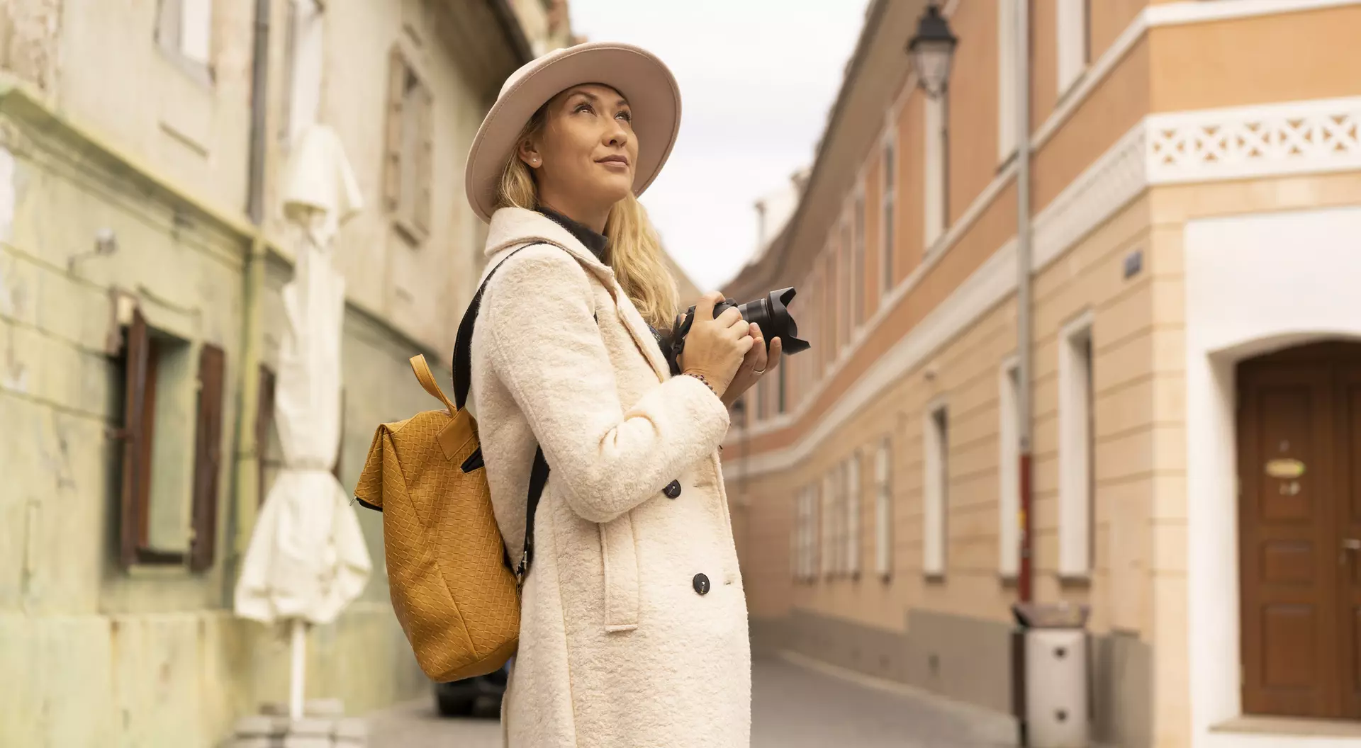 A woman with a camera looks up while standing in a charming, narrow street with colorful buildings.