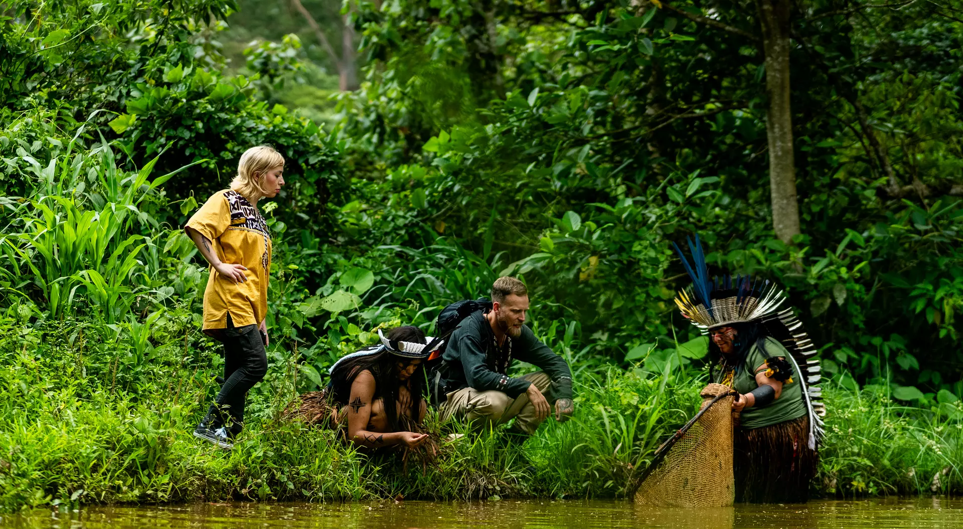 Three people interact by a river in a lush forest, one in traditional attire paddling a canoe, with two others observing.