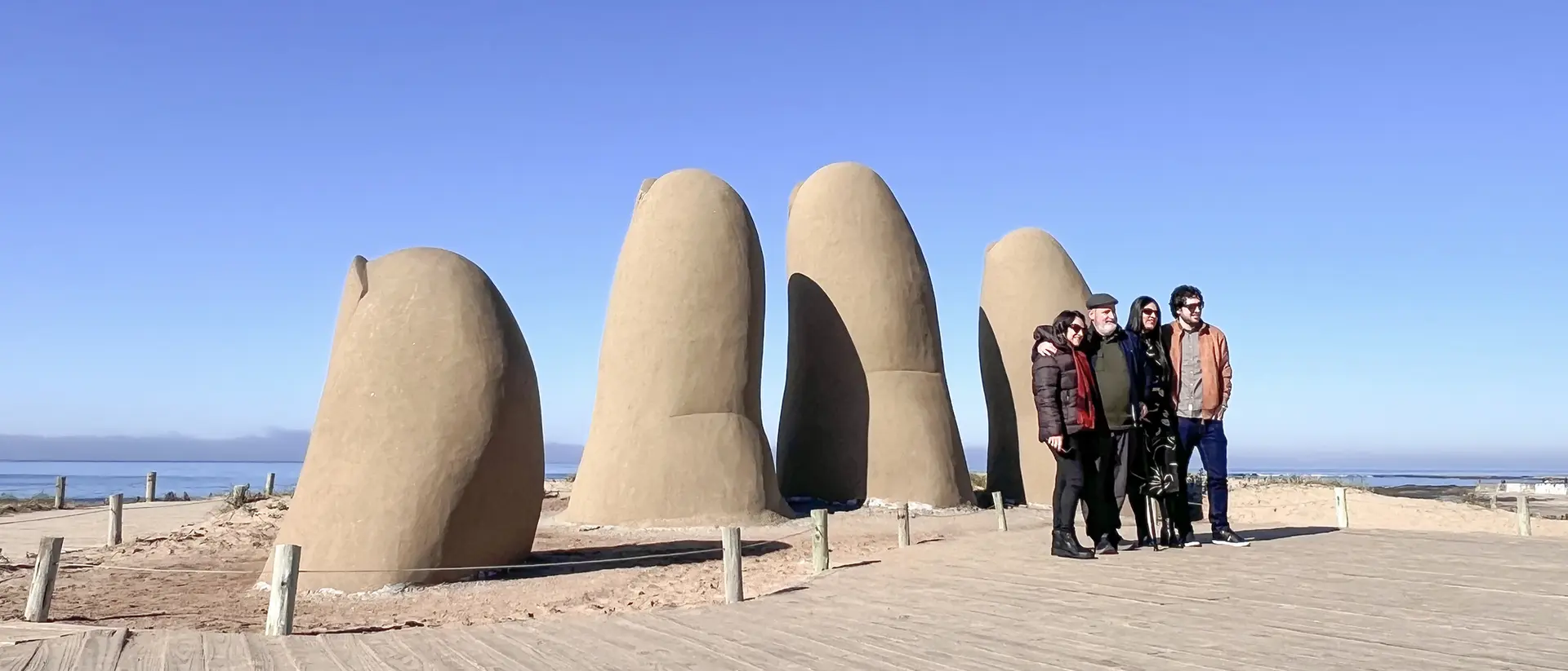 A group of six people stands in front of large, sculpted fingers on a sandy path with a clear blue sky.