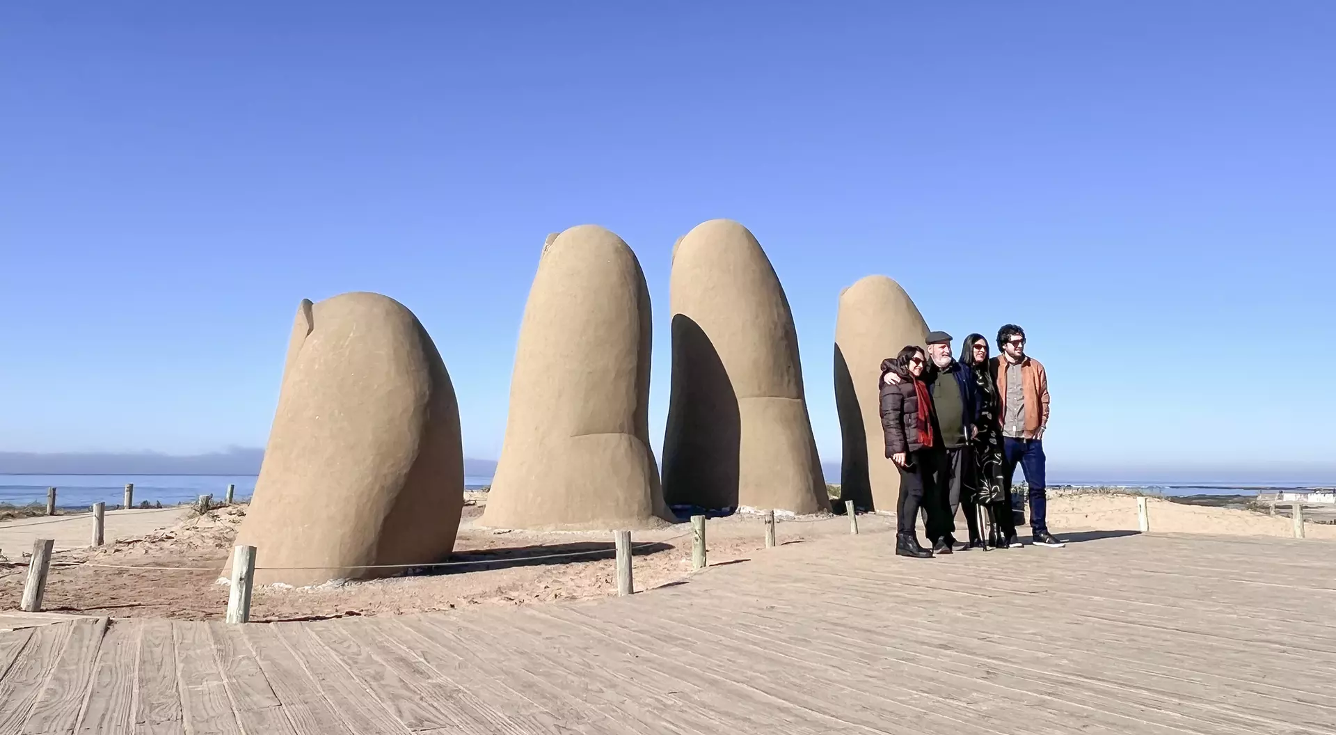 A group of six people stands in front of large, sculpted fingers on a sandy path with a clear blue sky.