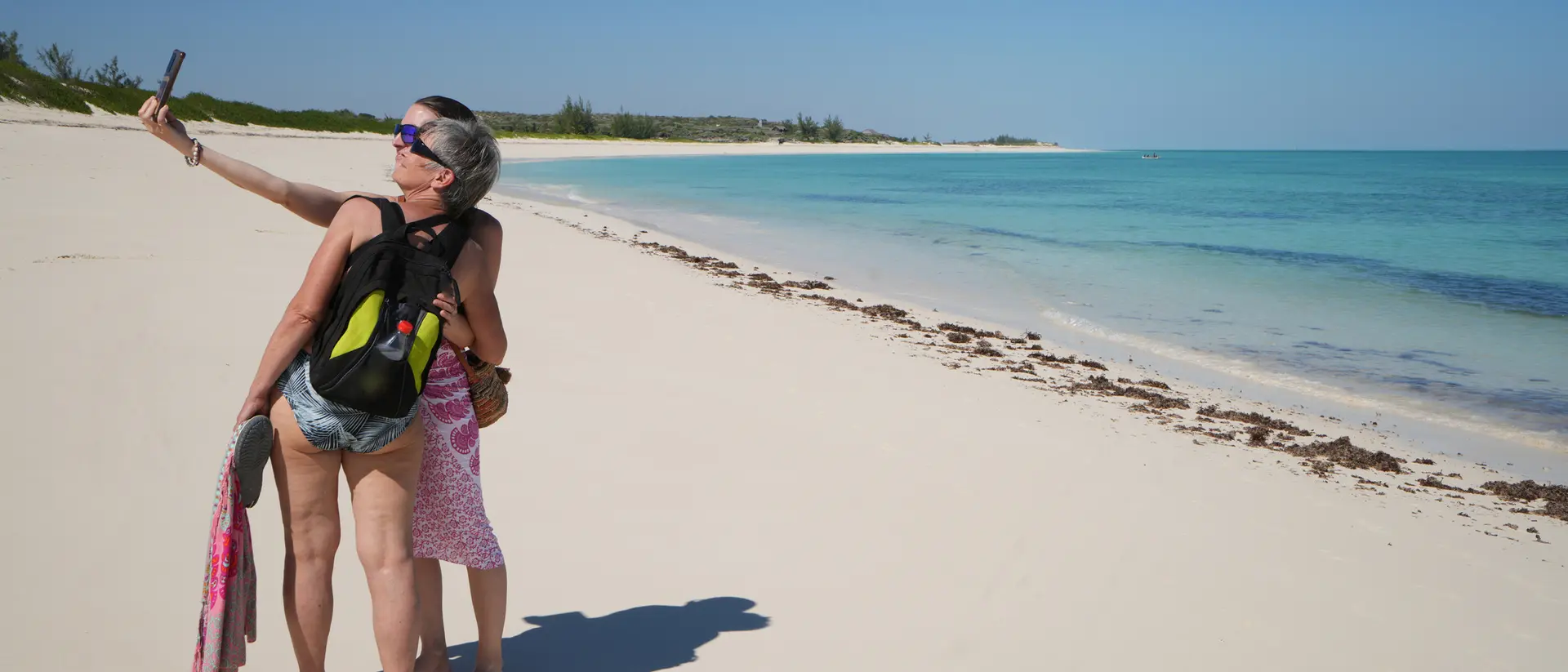 Two people take a selfie on a sunny beach with turquoise water and white sand in the background.