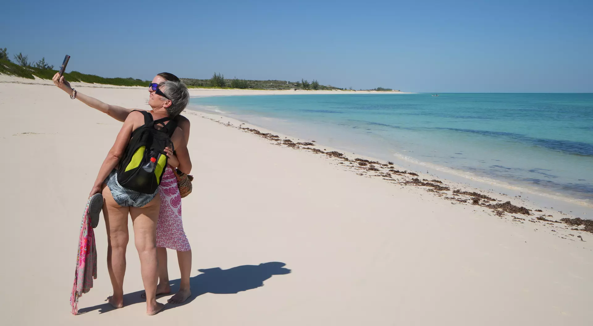 Two people take a selfie on a sunny beach with turquoise water and white sand in the background.