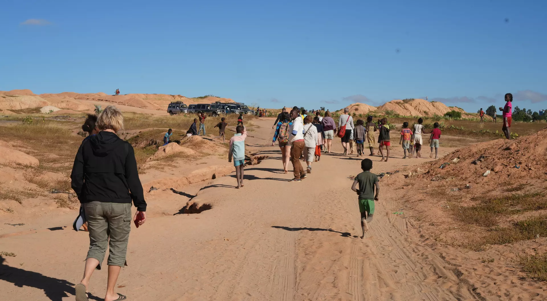 A group of people walking on a sandy path, with hills in the background and a clear blue sky. Kids play nearby.
