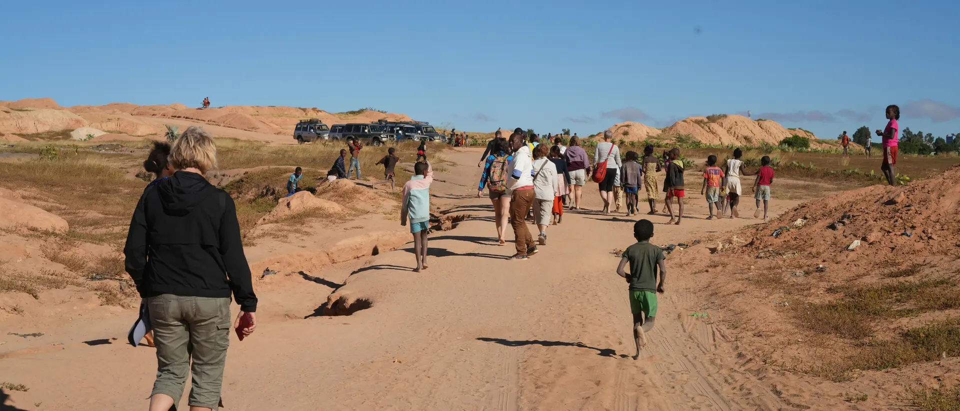 A group of people walking on a sandy path, with hills in the background and a clear blue sky. Kids play nearby.