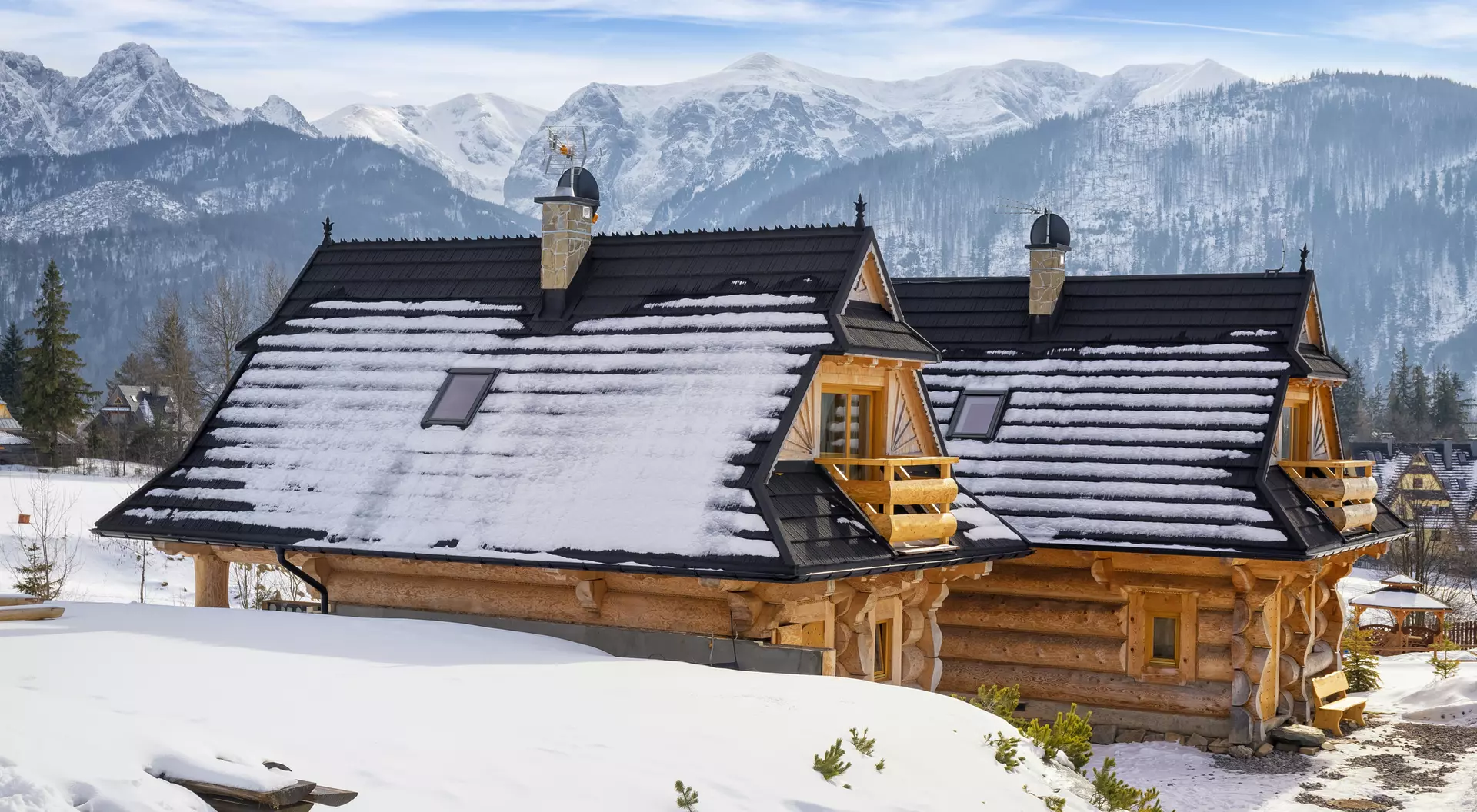 Two log cabins covered in snow, set against a backdrop of mountains under a clear blue sky.