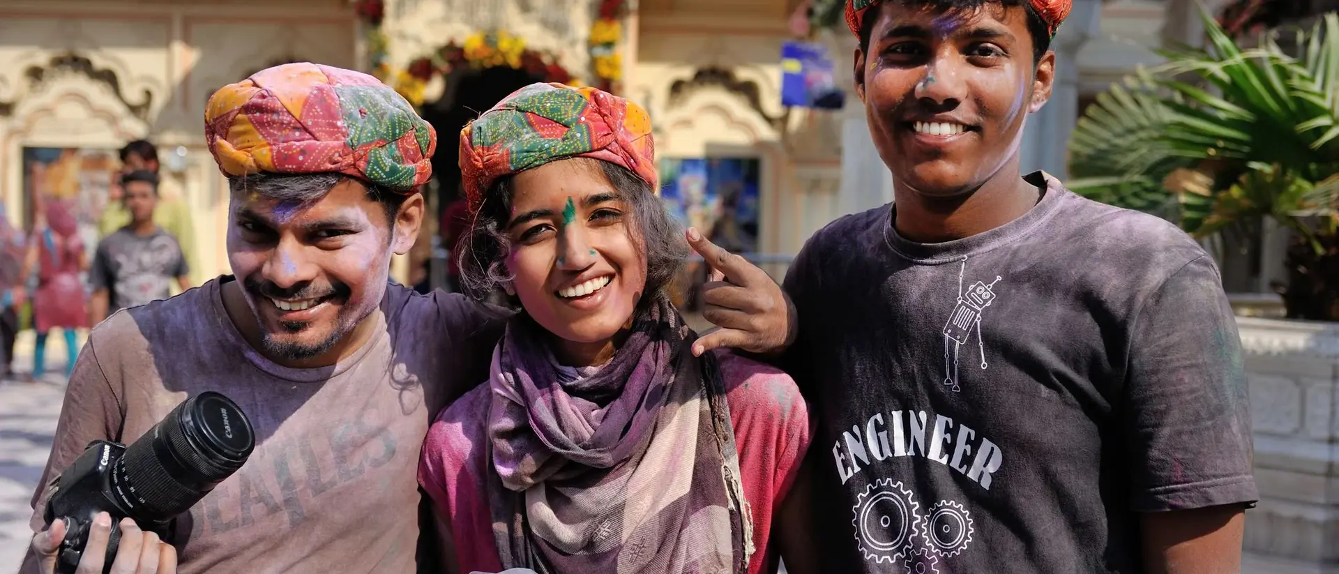 Three smiling friends in colorful turbans, holding cameras, pose together in a festive outdoor setting.