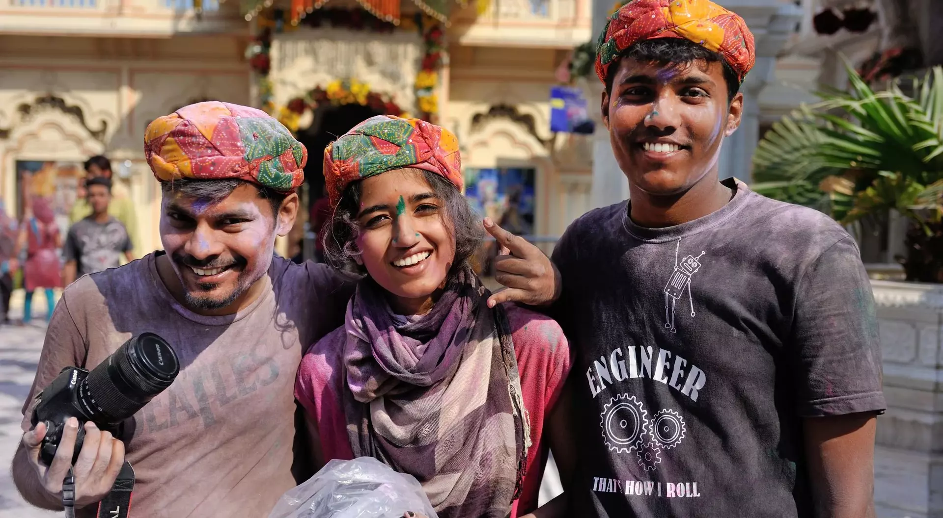 Three smiling friends in colorful turbans, holding cameras, pose together in a festive outdoor setting.