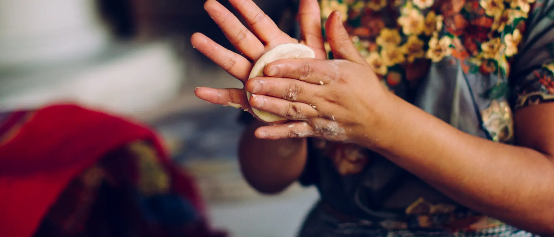 A person is shaping dough in their hands, wearing a colorful floral top with a checkered apron.