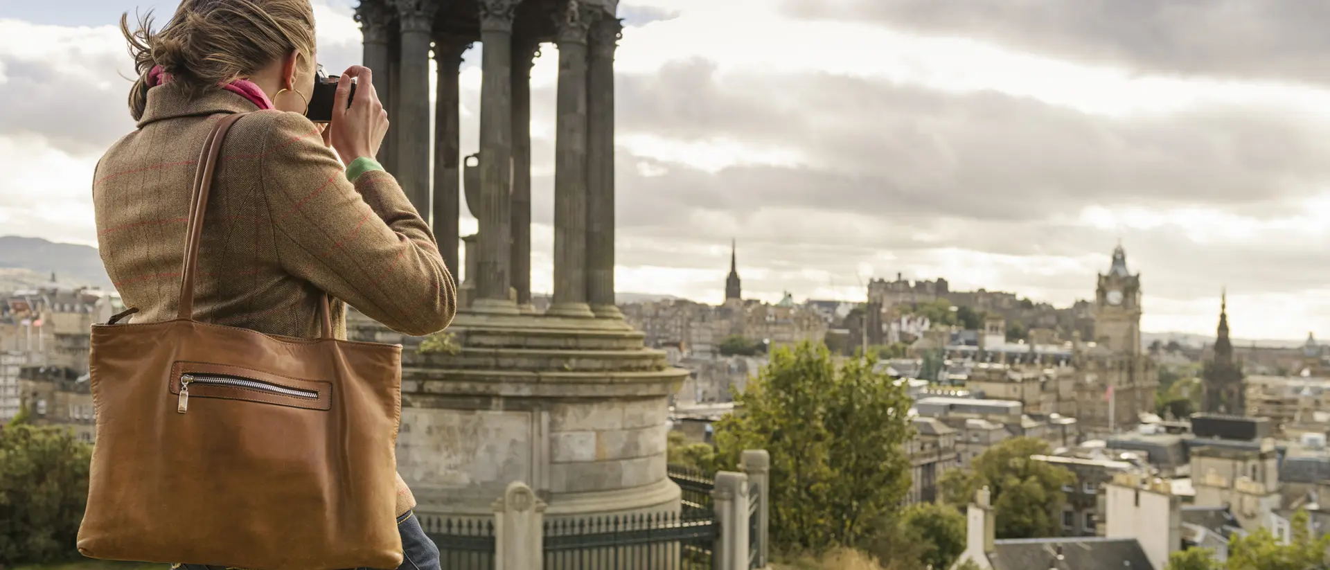 A person with a brown bag takes a photo of a cityscape featuring a monument against a cloudy sky.