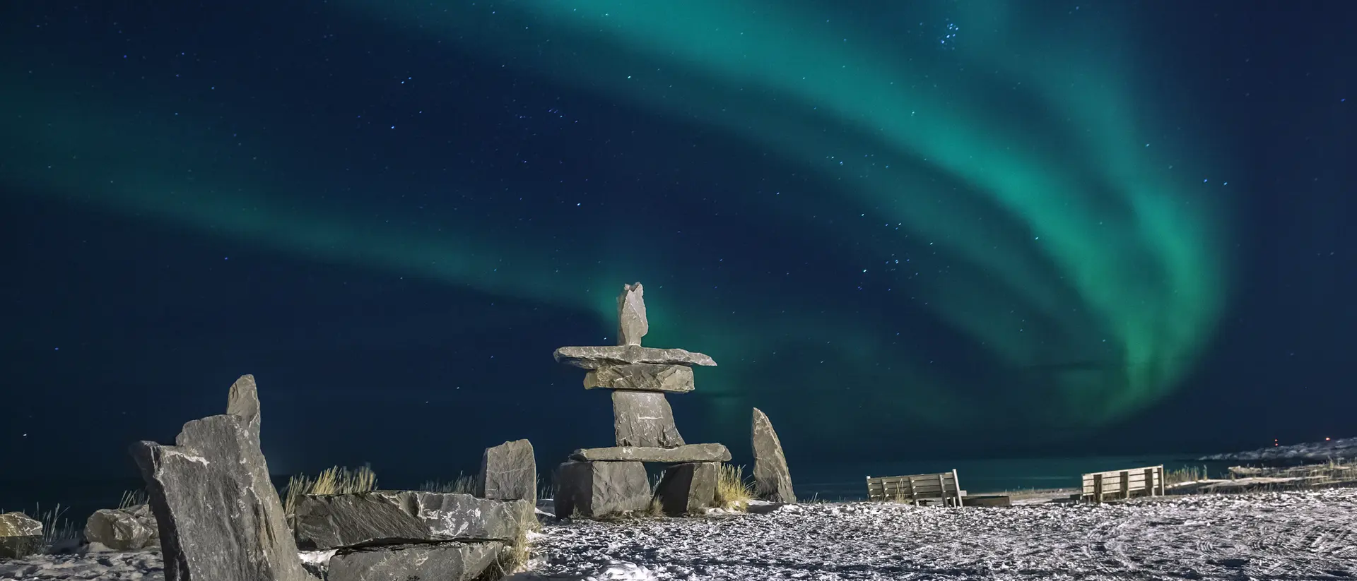 Inuit stone structure surrounded by snow, with vibrant green northern lights illuminating the night sky.