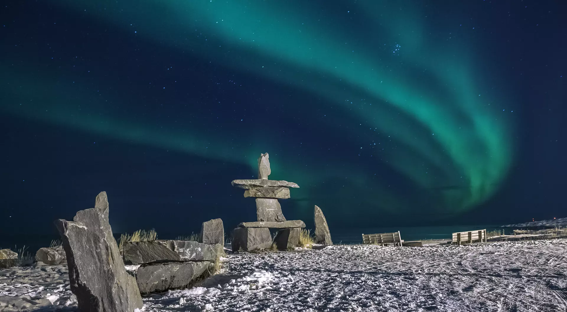 Inuit stone structure surrounded by snow, with vibrant green northern lights illuminating the night sky.