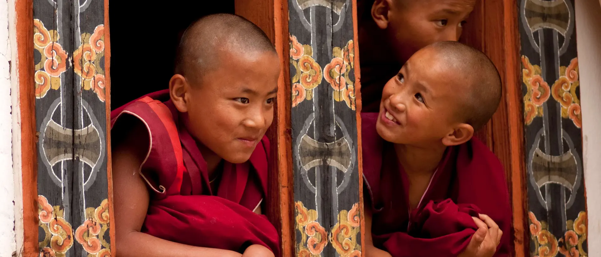 Three young monks with shaved heads peek out from a beautifully decorated window, smiling and engaging with each other.