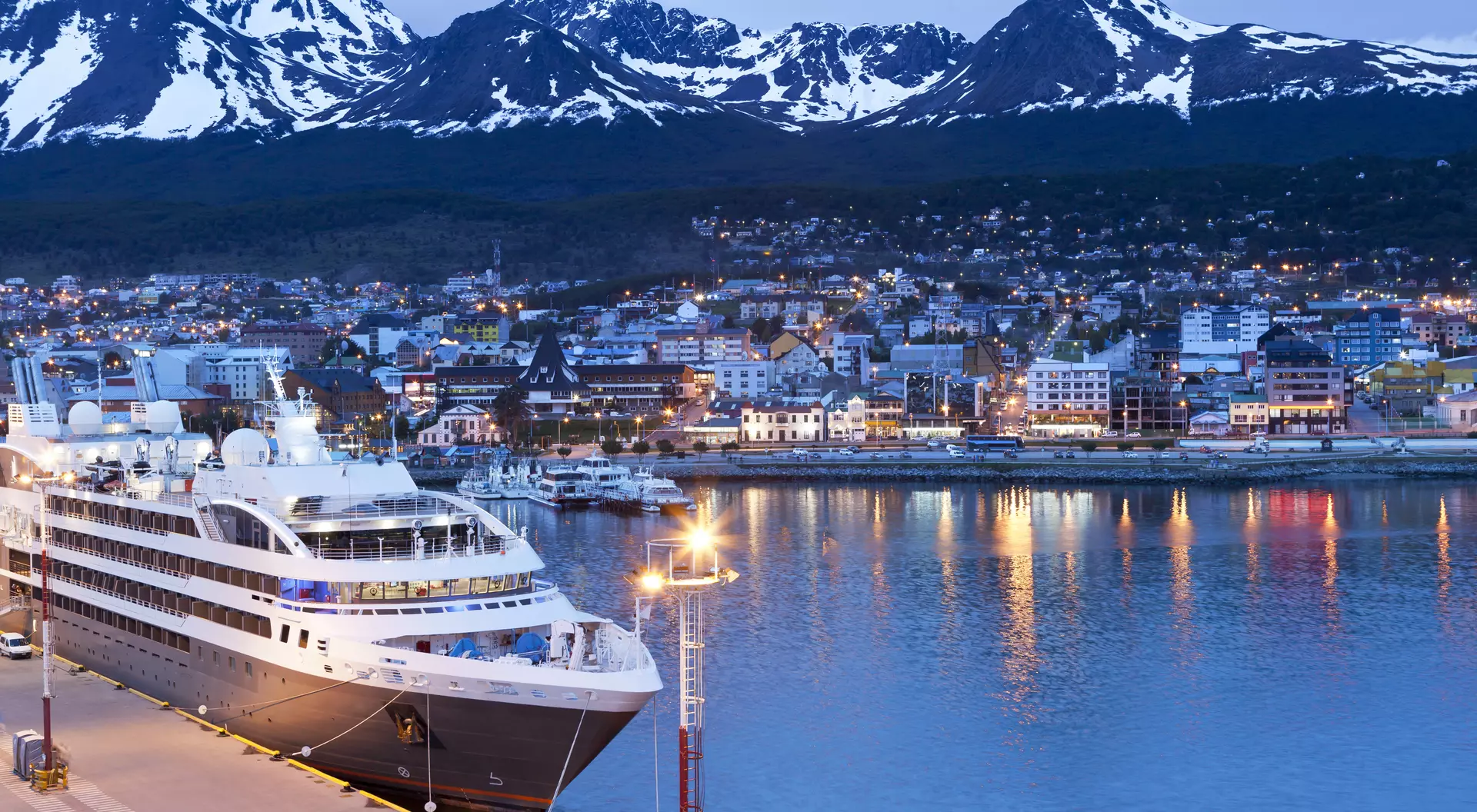 A cruise ship docked by a city harbor, surrounded by snow-capped mountains at dusk, with city lights reflecting on the water.