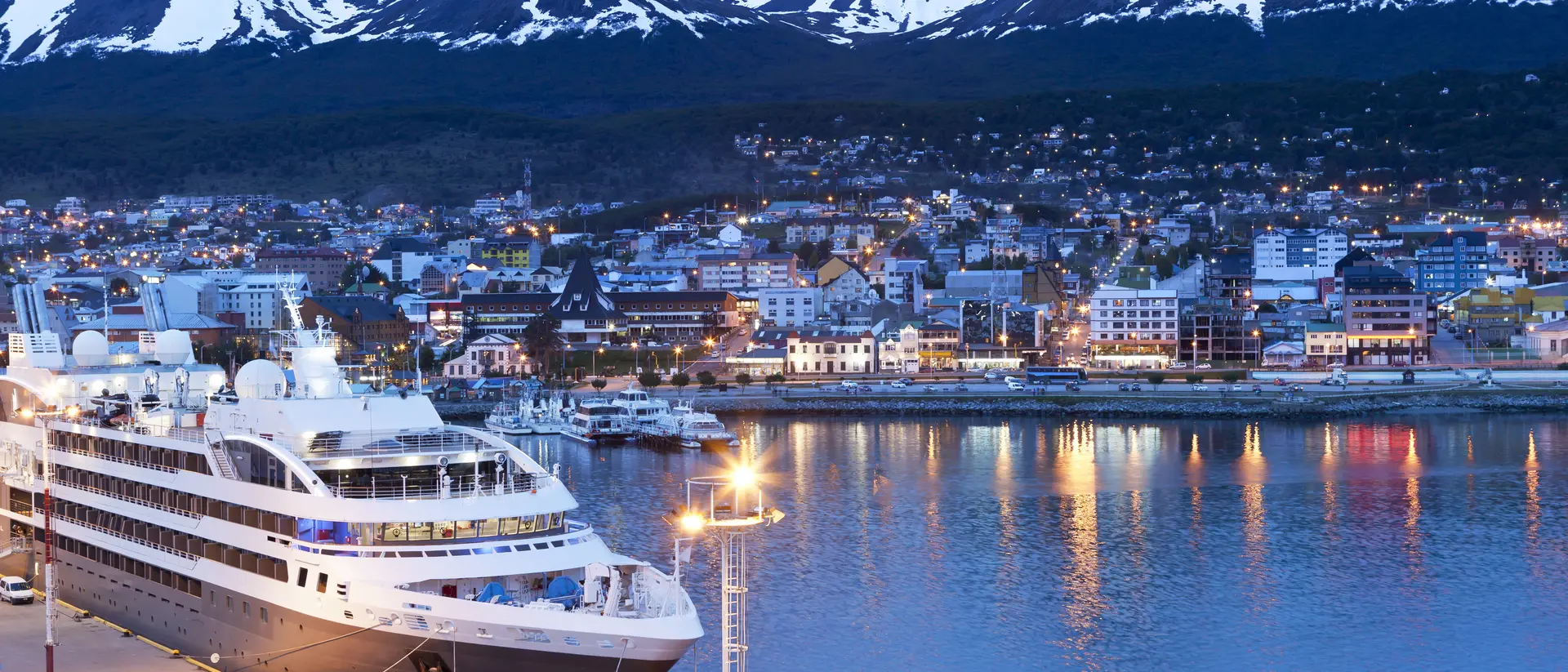 A cruise ship docked by a city harbor, surrounded by snow-capped mountains at dusk, with city lights reflecting on the water.