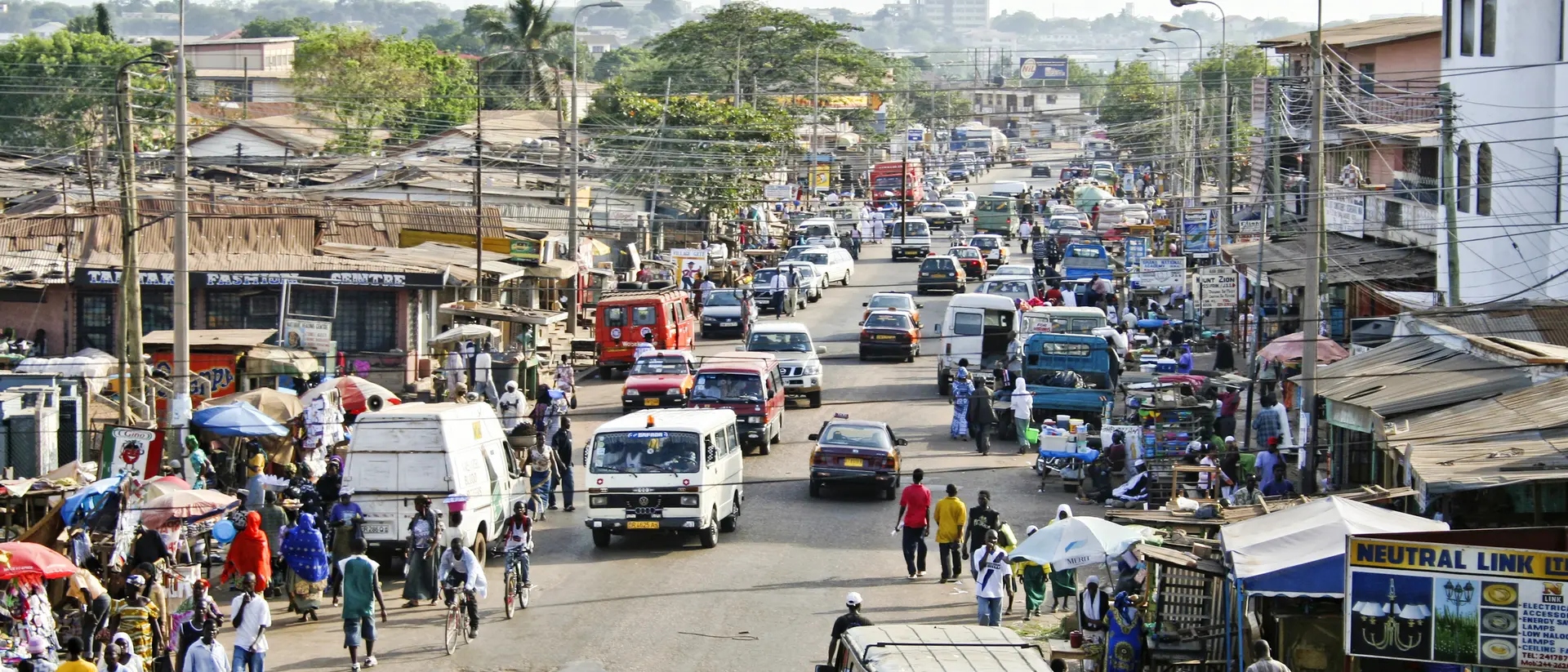 Busy street scene in a town, filled with vehicles, vendors, and pedestrians amidst shops and buildings.