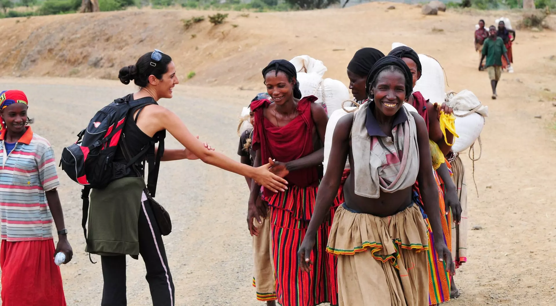 A person greets a group of women in colorful clothing on a dirt road, with mountains in the background.