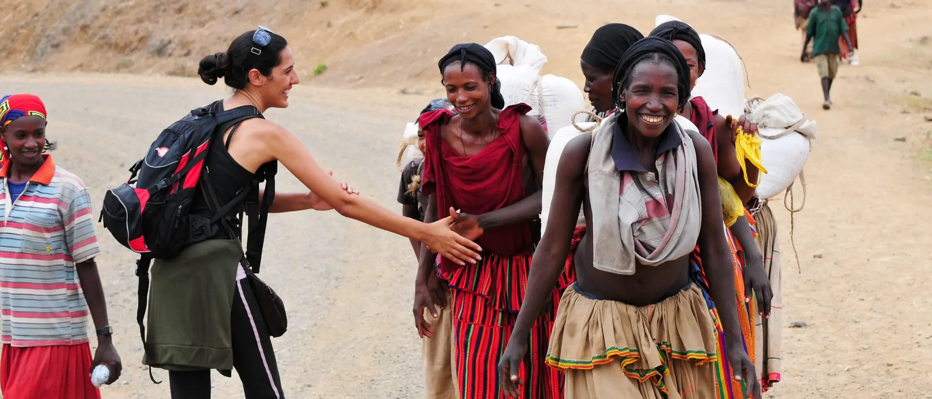 A person greets a group of women in colorful clothing on a dirt road, with mountains in the background.