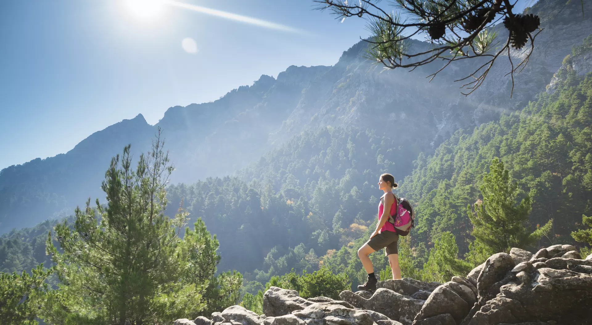 A hiker stands on rocky terrain, surrounded by trees, mountains, and sunlight filtering through the landscape.