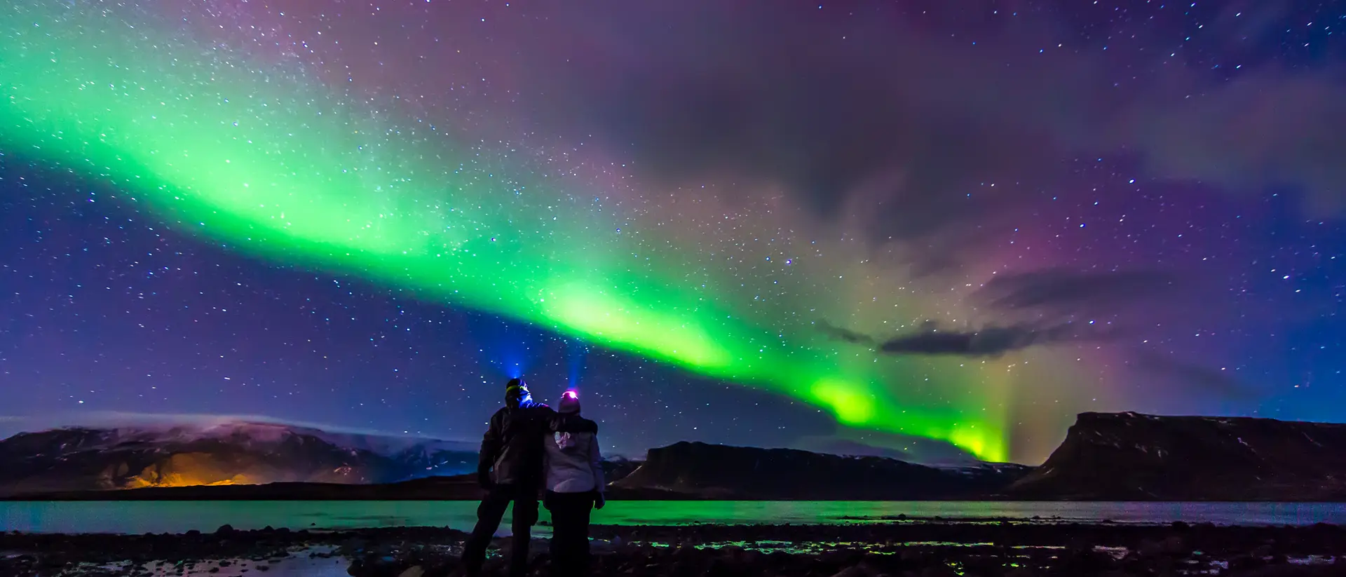 Two people stand on a rocky shore, gazing at the vibrant green northern lights illuminating the night sky.