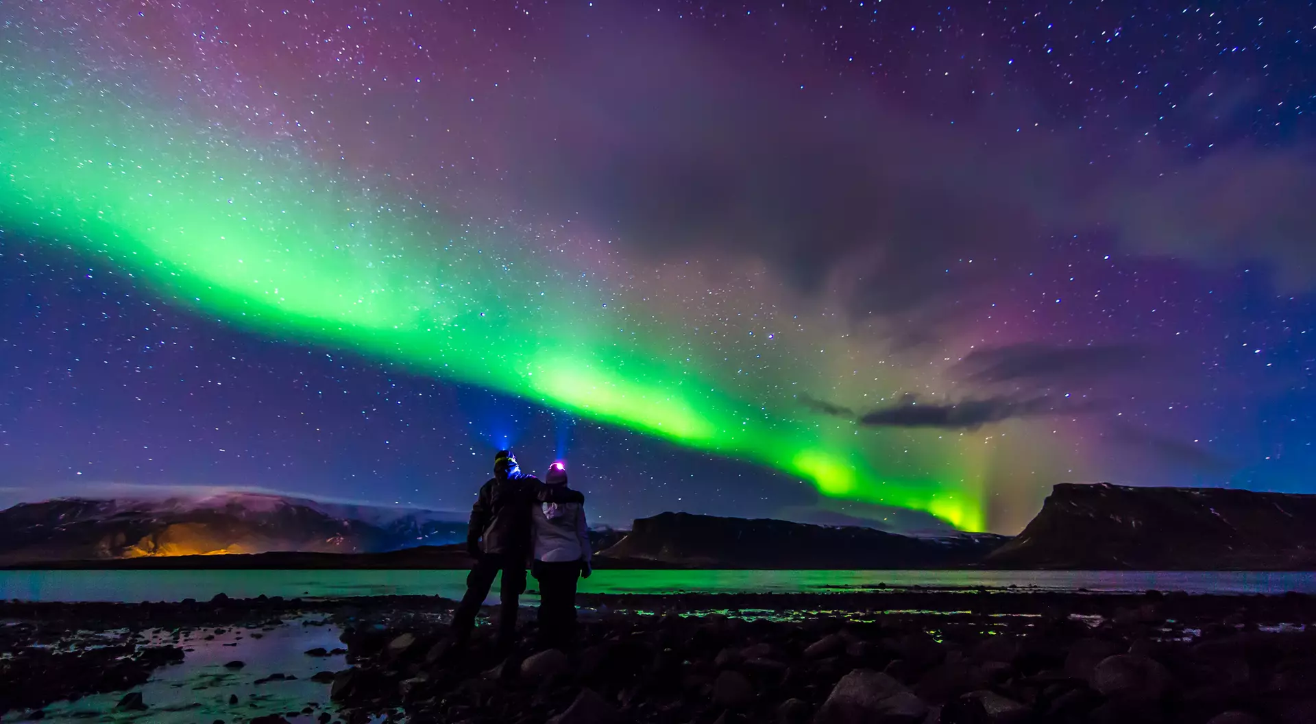 Two people stand on a rocky shore, gazing at the vibrant green northern lights illuminating the night sky.
