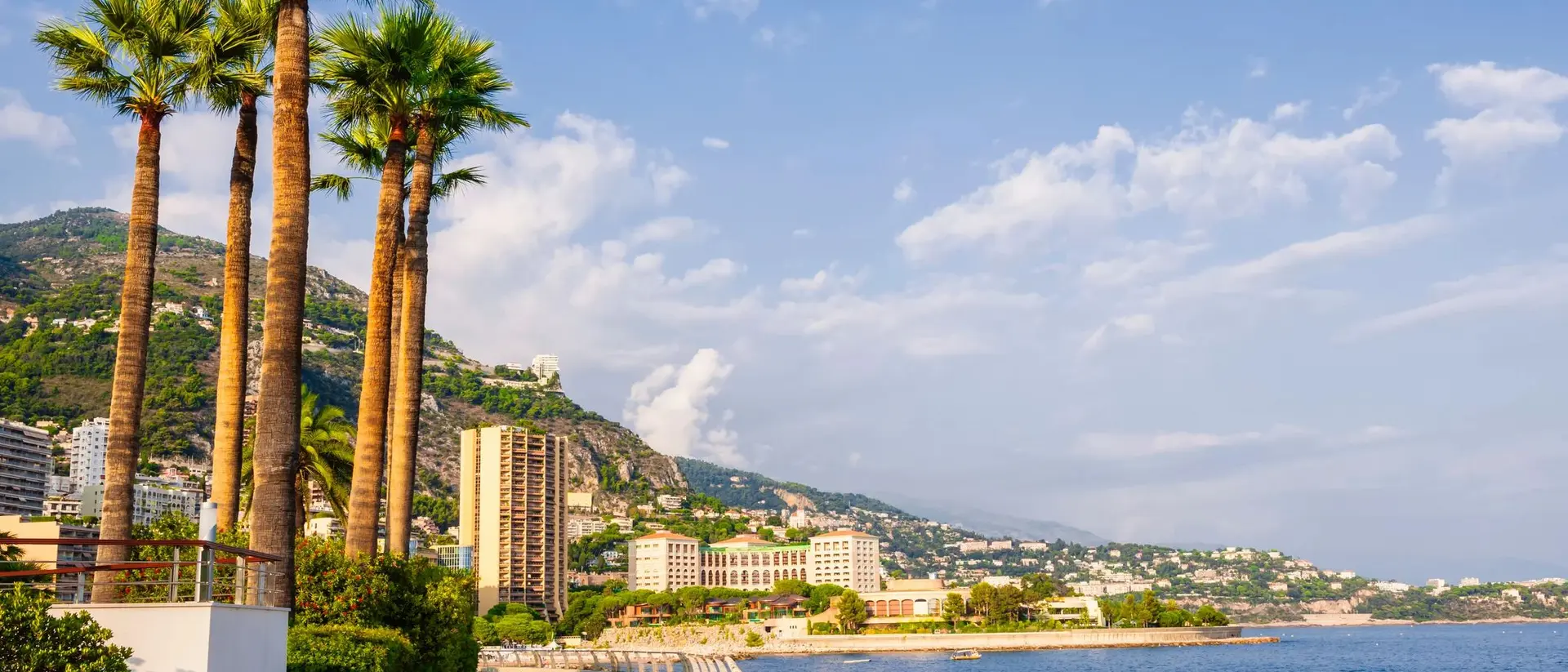 Scenic view of a coastal promenade lined with palm trees, buildings, and mountains under a blue sky with clouds.