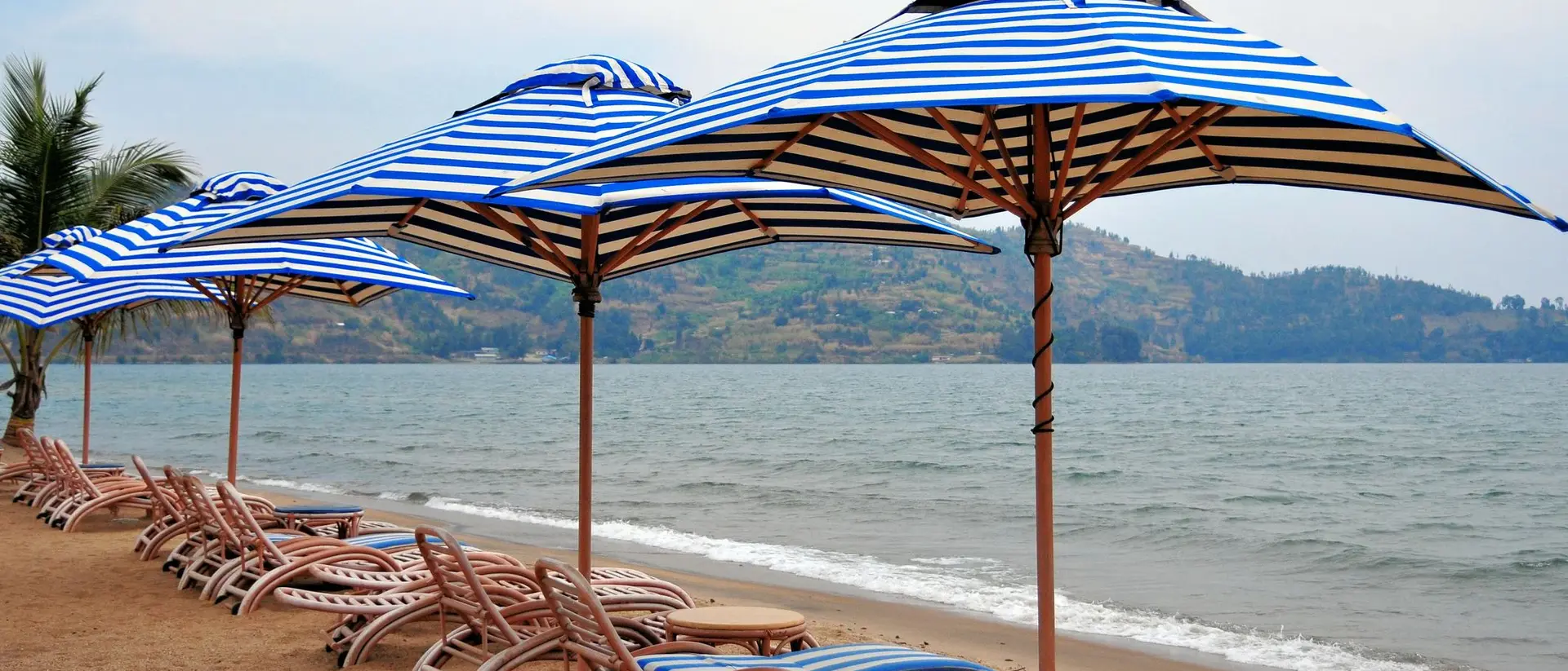 Row of blue and white striped beach umbrellas on sandy shore, with lounge chairs and calm sea in the background.