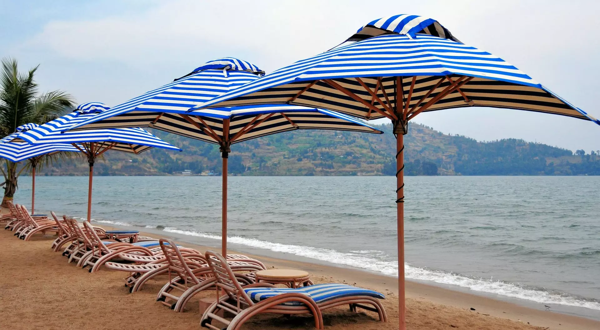 Row of blue and white striped beach umbrellas on sandy shore, with lounge chairs and calm sea in the background.