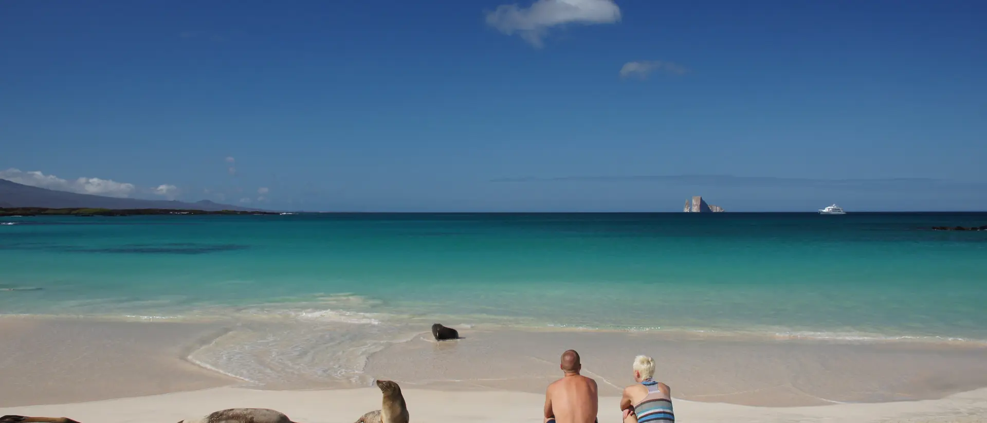 A couple sits on a sandy beach, gazing at the turquoise ocean and distant rock formations under a clear blue sky.