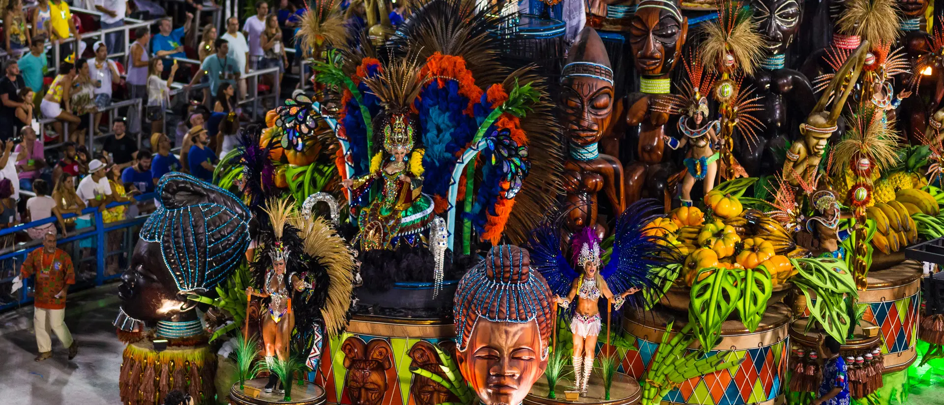 Colorful parade float featuring intricate masks and tropical motifs, surrounded by a crowd in vibrant attire.