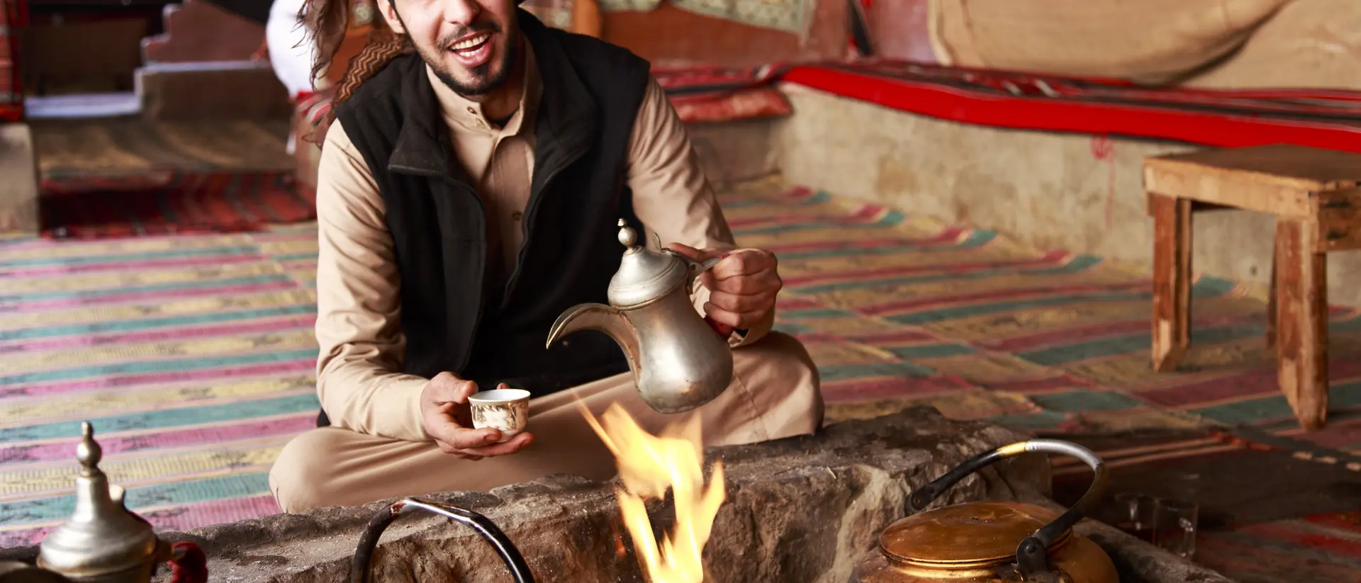 A man in traditional attire sits by a fire, pouring tea from a kettle in a cozy, tent-like setting.