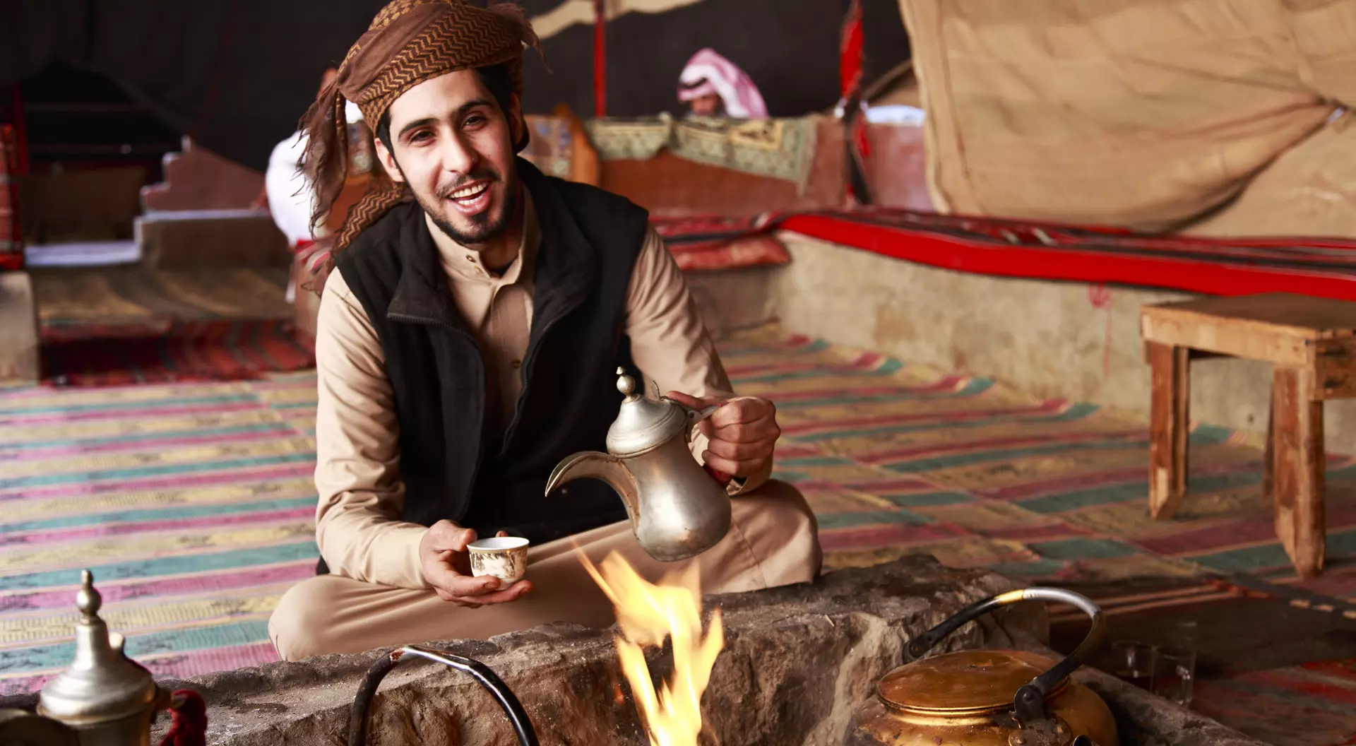 A man in traditional attire sits by a fire, pouring tea from a kettle in a cozy, tent-like setting.