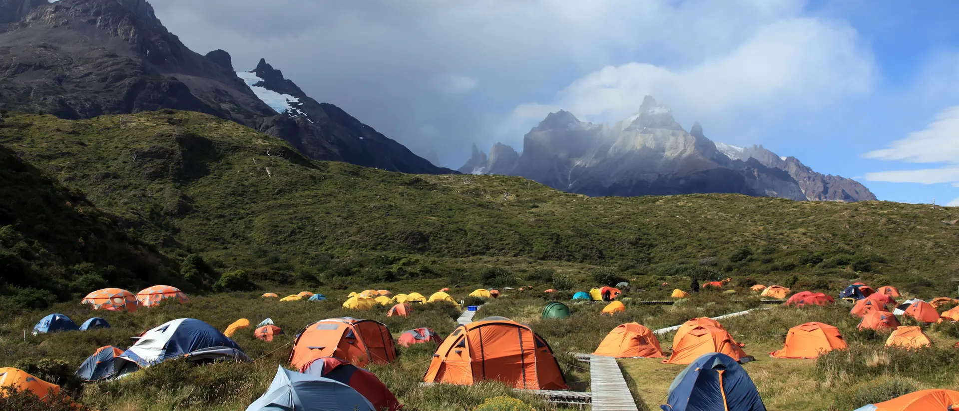 A campsite with colorful tents set in a lush valley, framed by rugged mountains and a partly cloudy sky.