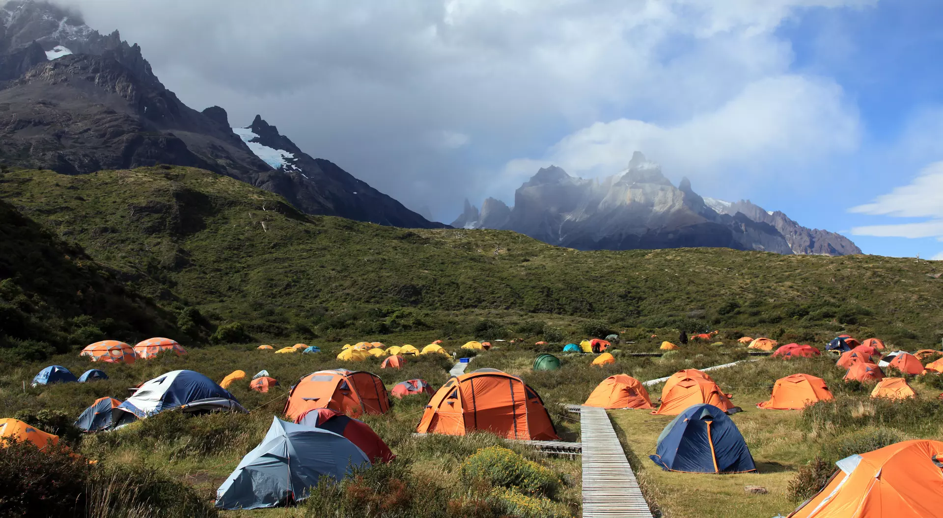 A campsite with colorful tents set in a lush valley, framed by rugged mountains and a partly cloudy sky.