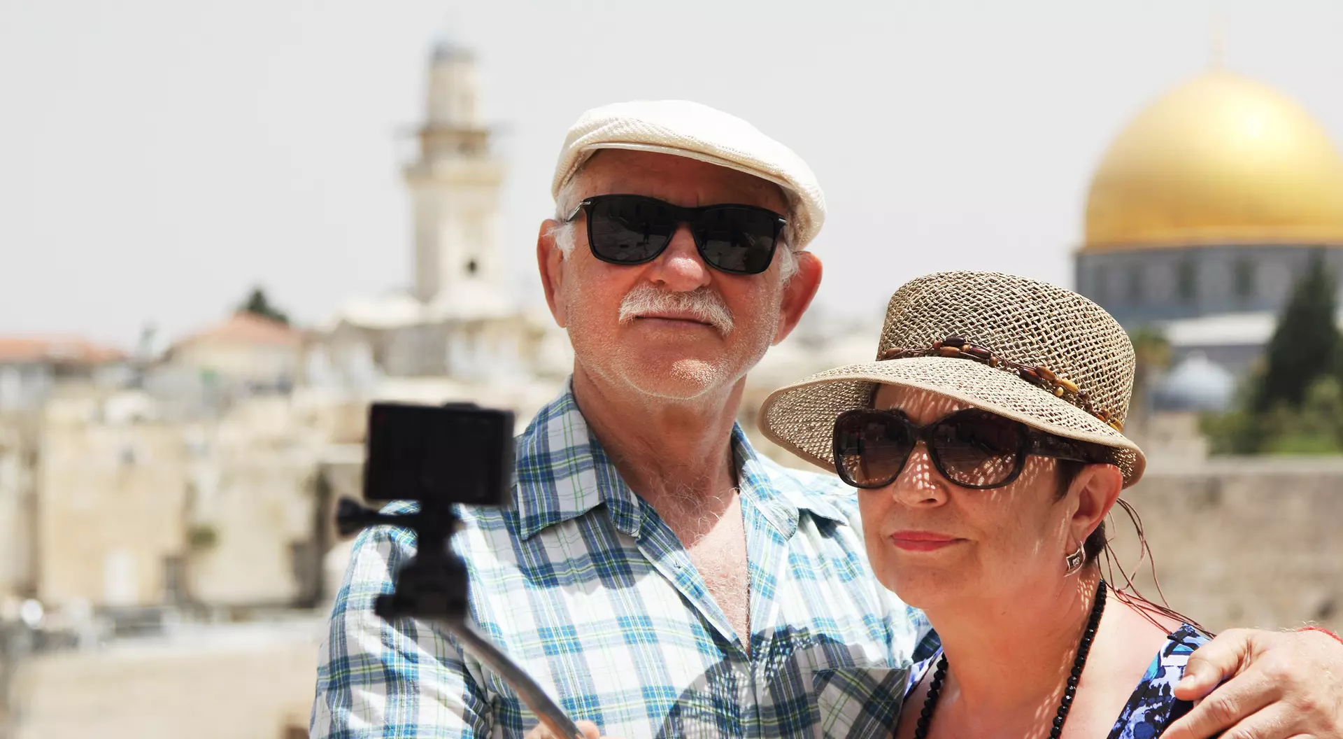 An older couple taking a selfie with a cityscape and a golden dome in the background on a sunny day.