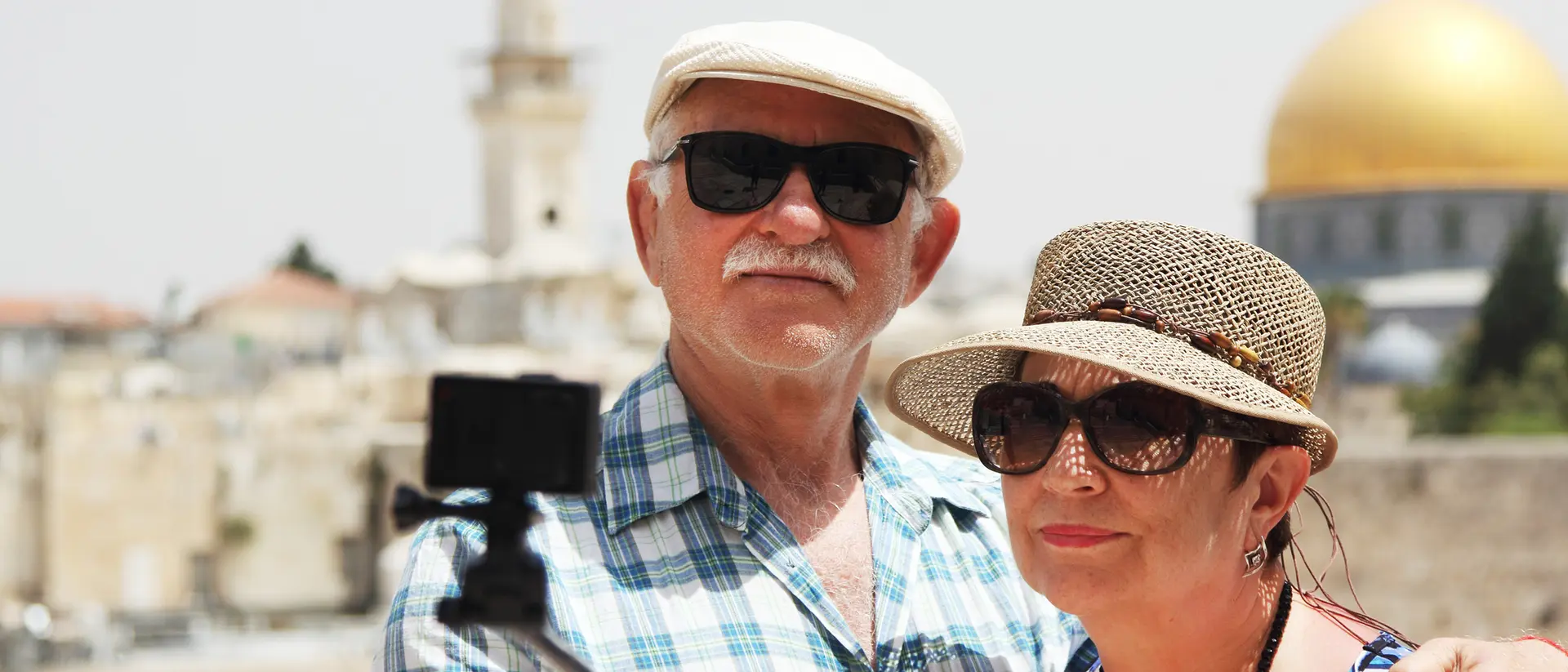 An older couple taking a selfie with a cityscape and a golden dome in the background on a sunny day.