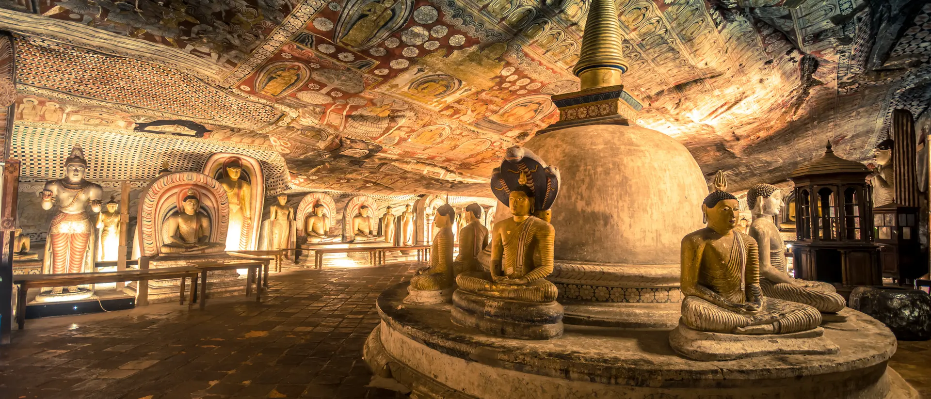 Interior of a cave temple featuring ornate frescoes, seated Buddha statues, and a central stupa under a colorful ceiling.