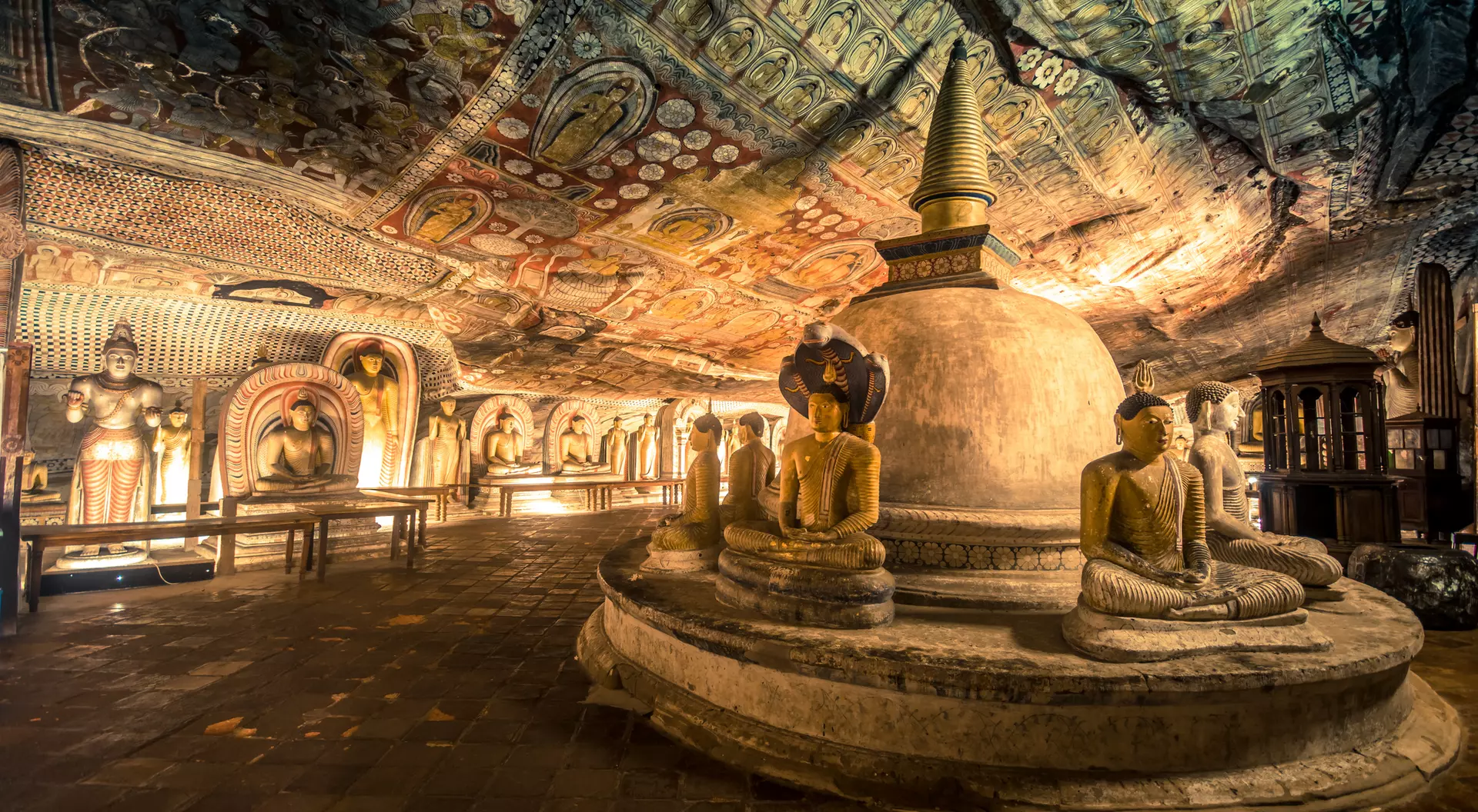 Interior of a cave temple featuring ornate frescoes, seated Buddha statues, and a central stupa under a colorful ceiling.