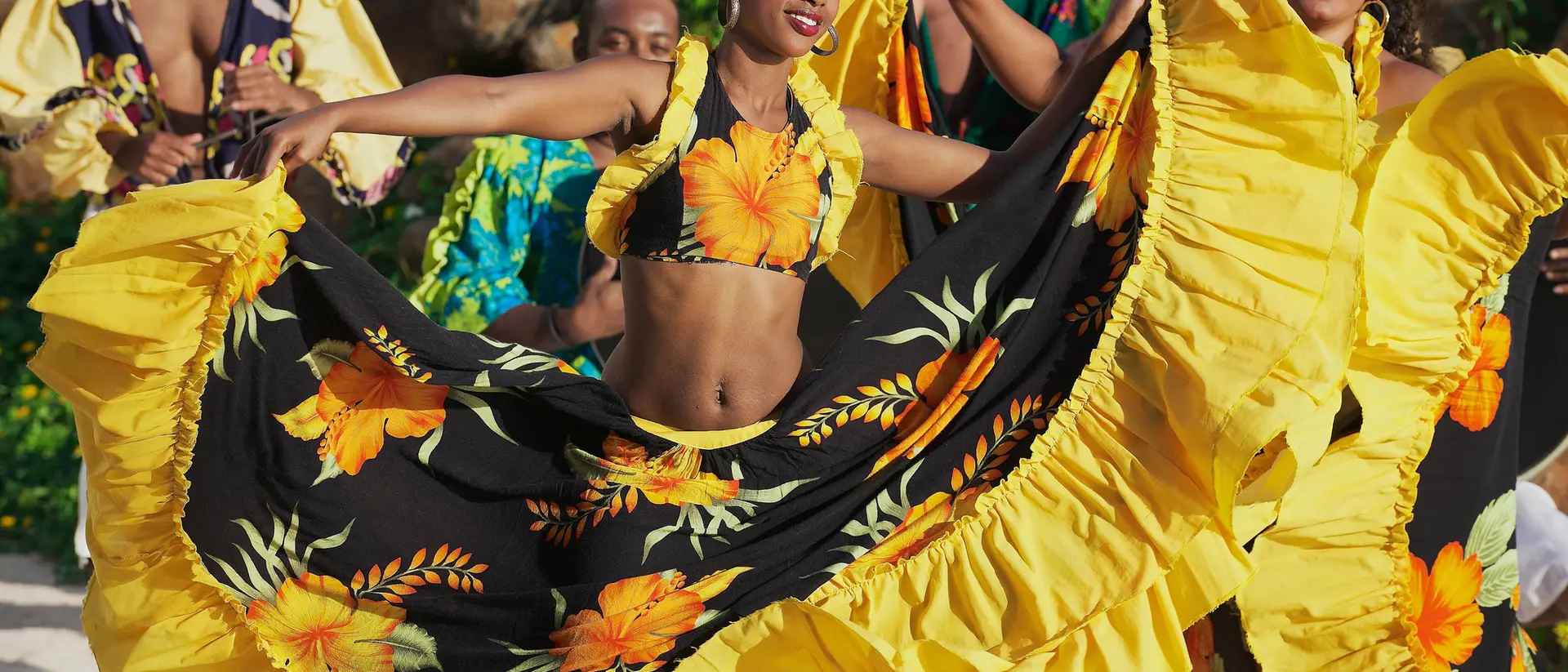 Two women in vibrant yellow and black dresses dance joyfully, surrounded by musicians dressed in colorful attire.