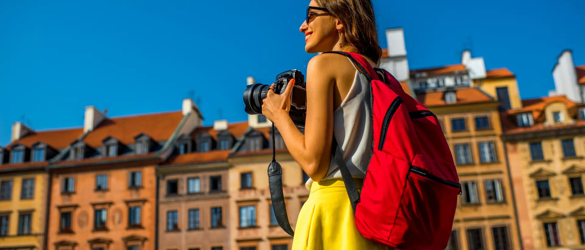 Smiling woman with a camera and red backpack stands in a sunny square with colorful buildings in the background.