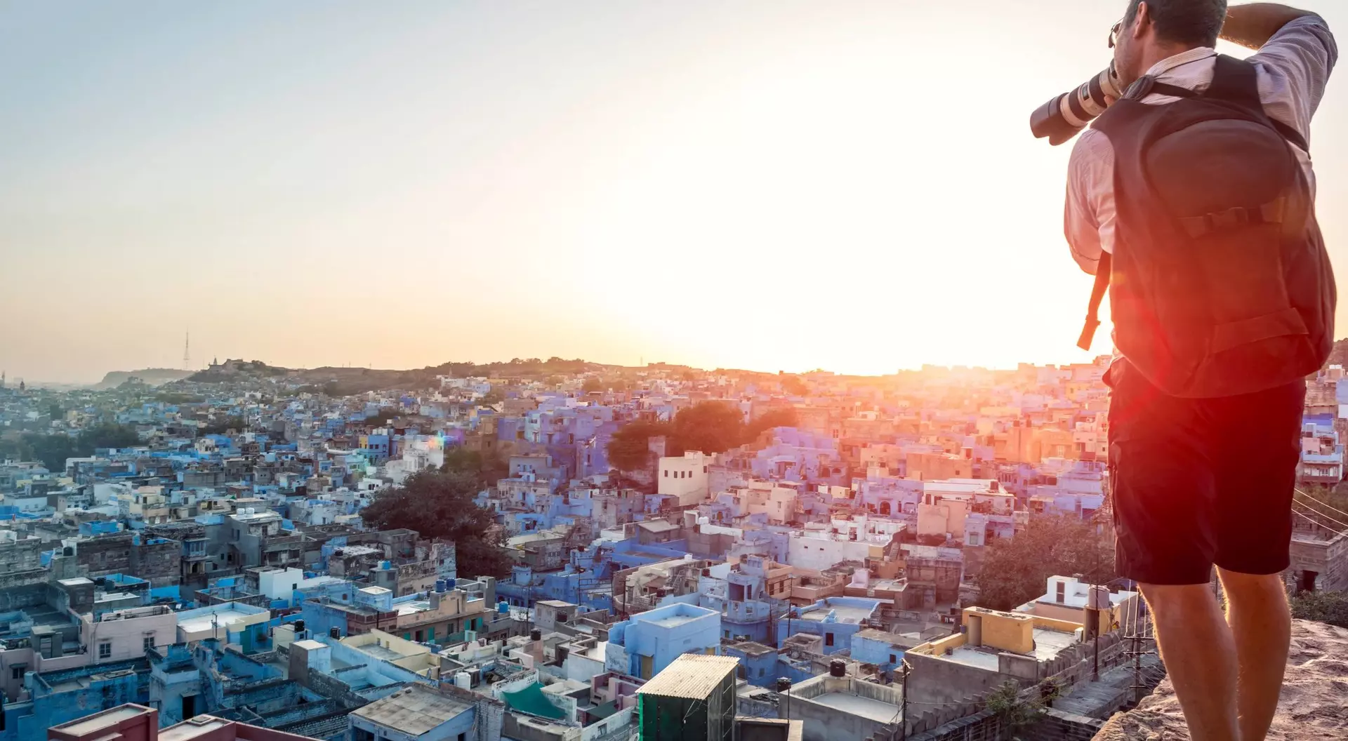 A photographer stands on a hilltop, capturing a sunset over a cityscape of blue buildings.