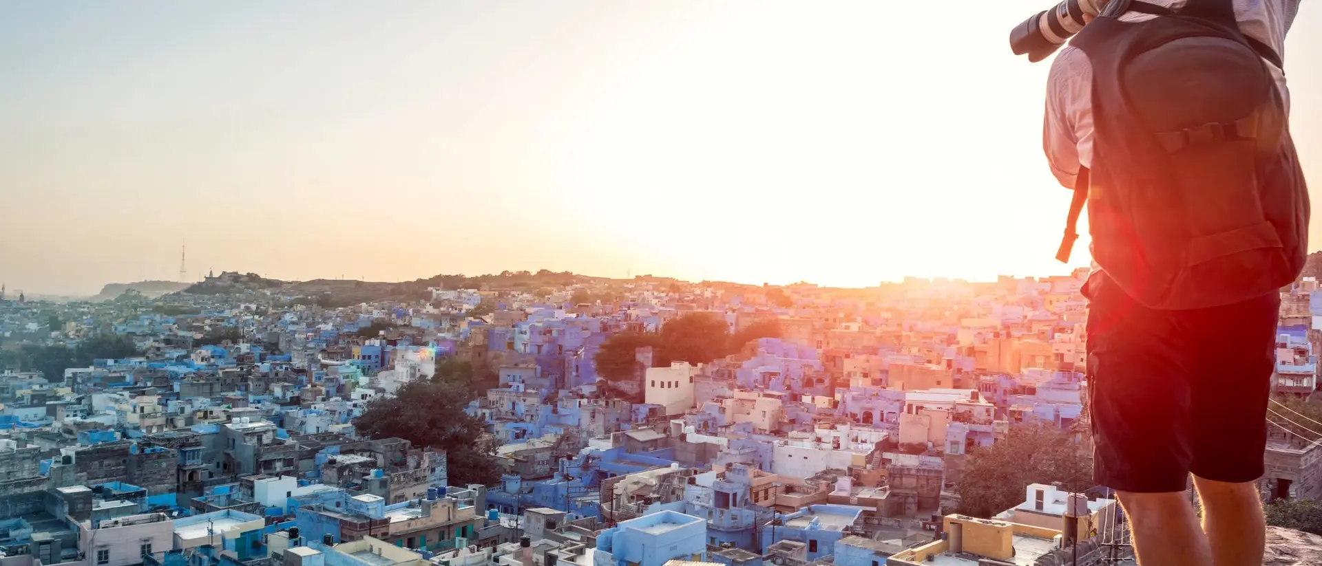 A photographer stands on a hilltop, capturing a sunset over a cityscape of blue buildings.