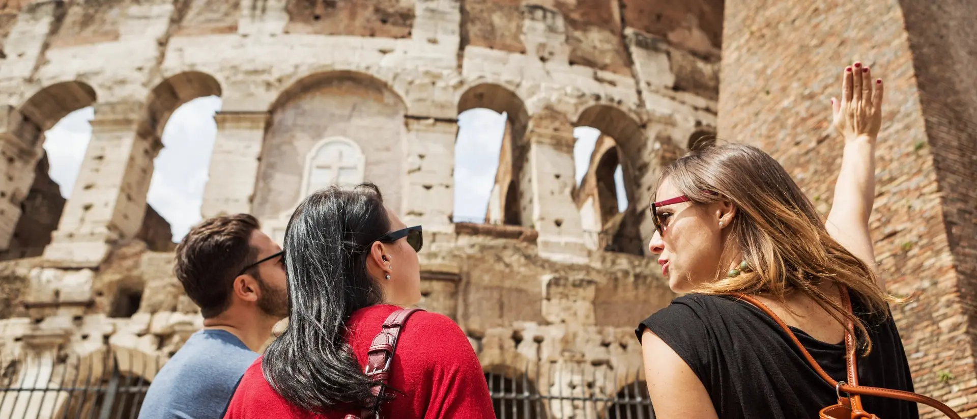 Three people admire the Colosseum, with one pointing and explaining, set against a clear blue sky.