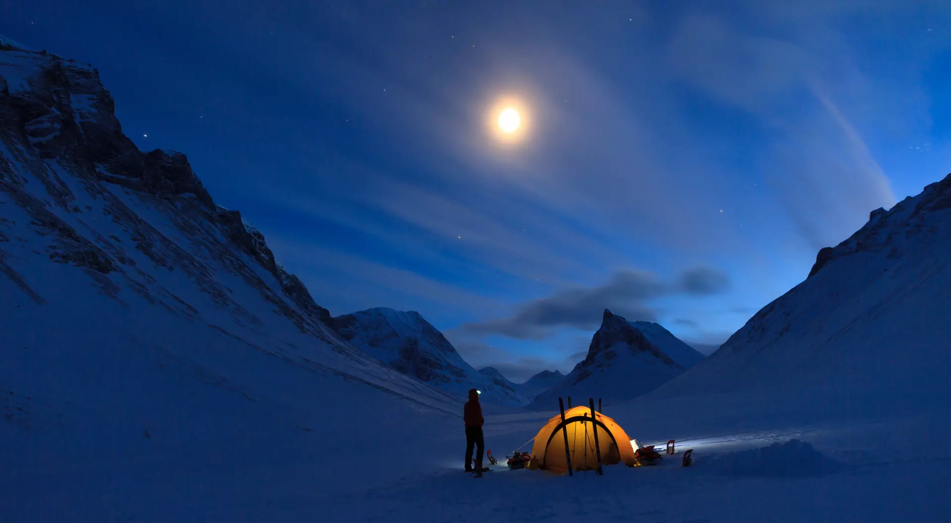 A glowing tent in a snowy valley under a moonlit sky, with mountains and clouds illuminated by the moonlight.