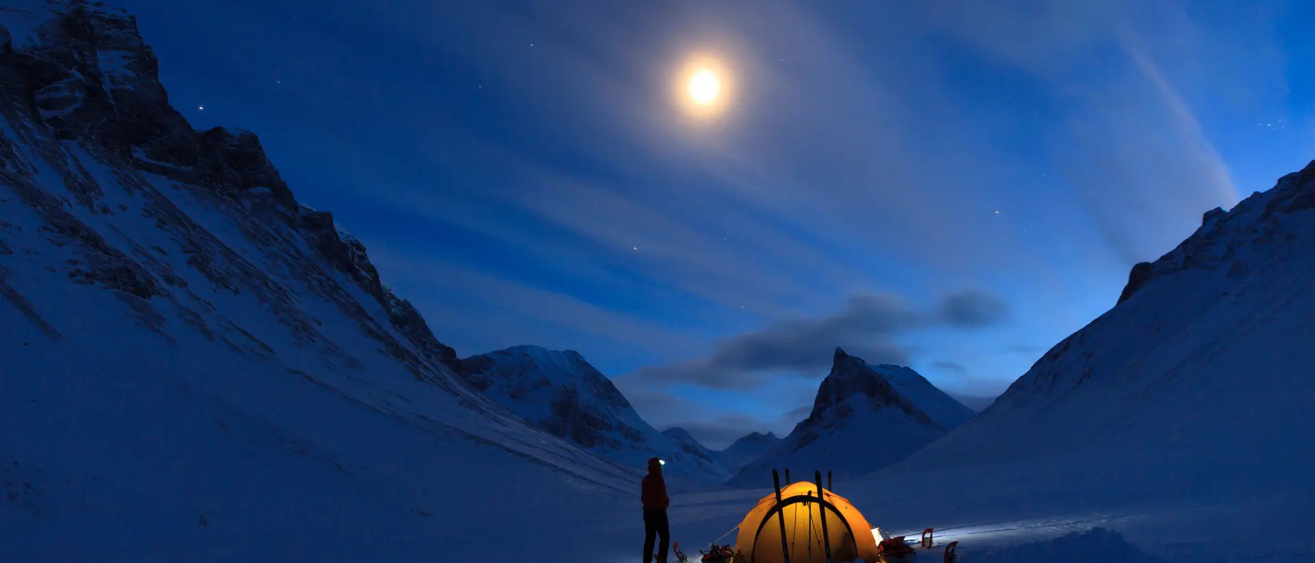 A glowing tent in a snowy valley under a moonlit sky, with mountains and clouds illuminated by the moonlight.