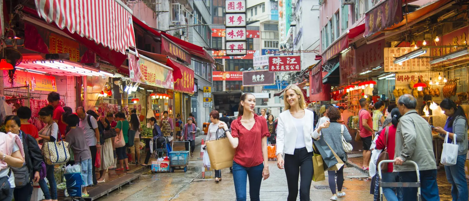 Two women walk through a bustling market street filled with colorful shops and people, vibrant signs in the background.