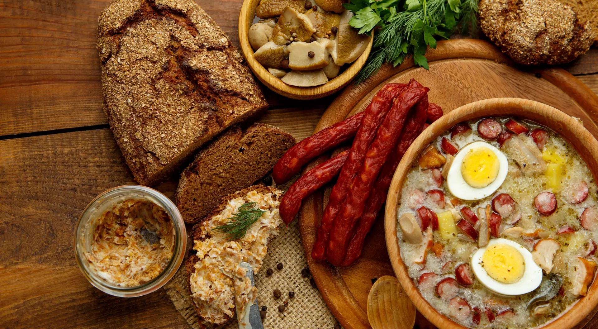 A rustic wooden table displays a hearty meal with soup, sliced sausage, bread, pickles, and garnishes.