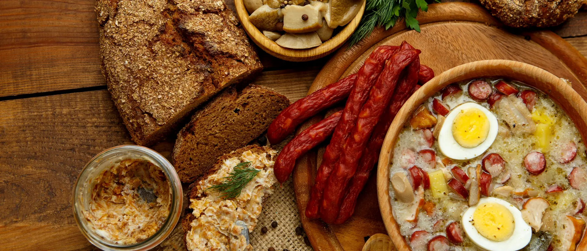A rustic wooden table displays a hearty meal with soup, sliced sausage, bread, pickles, and garnishes.
