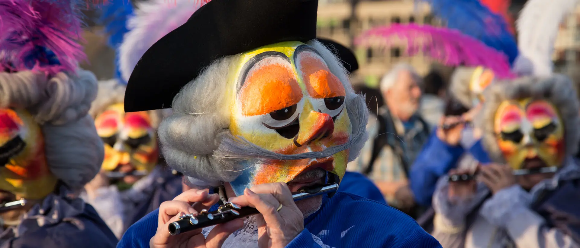 A performer in a feathered hat plays a flute, surrounded by others in colorful masks and costumes during a celebration.