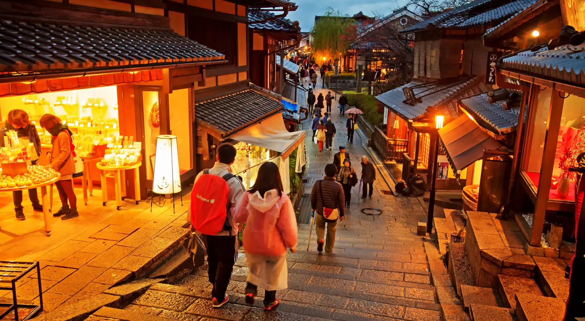 Two people walk down stone steps in a bustling street, lined with traditional shops and warm lighting during dusk.